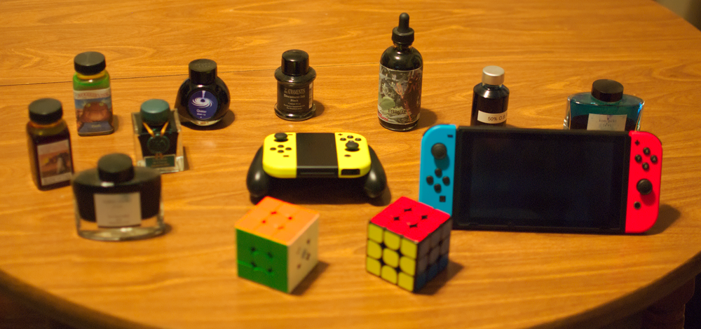 Various objects, a Nintendo Switch with blue / red joycons, a pair of yellow joycons, one rubik&rsquo;s cube with orange, dark green, and white showing, another with yellow, red, and blue showing, and various bottle of fountain pen ink, ranging from flourescent green, to purple, blue, black, and teal. The entire photo has a yellow tint from the white balance